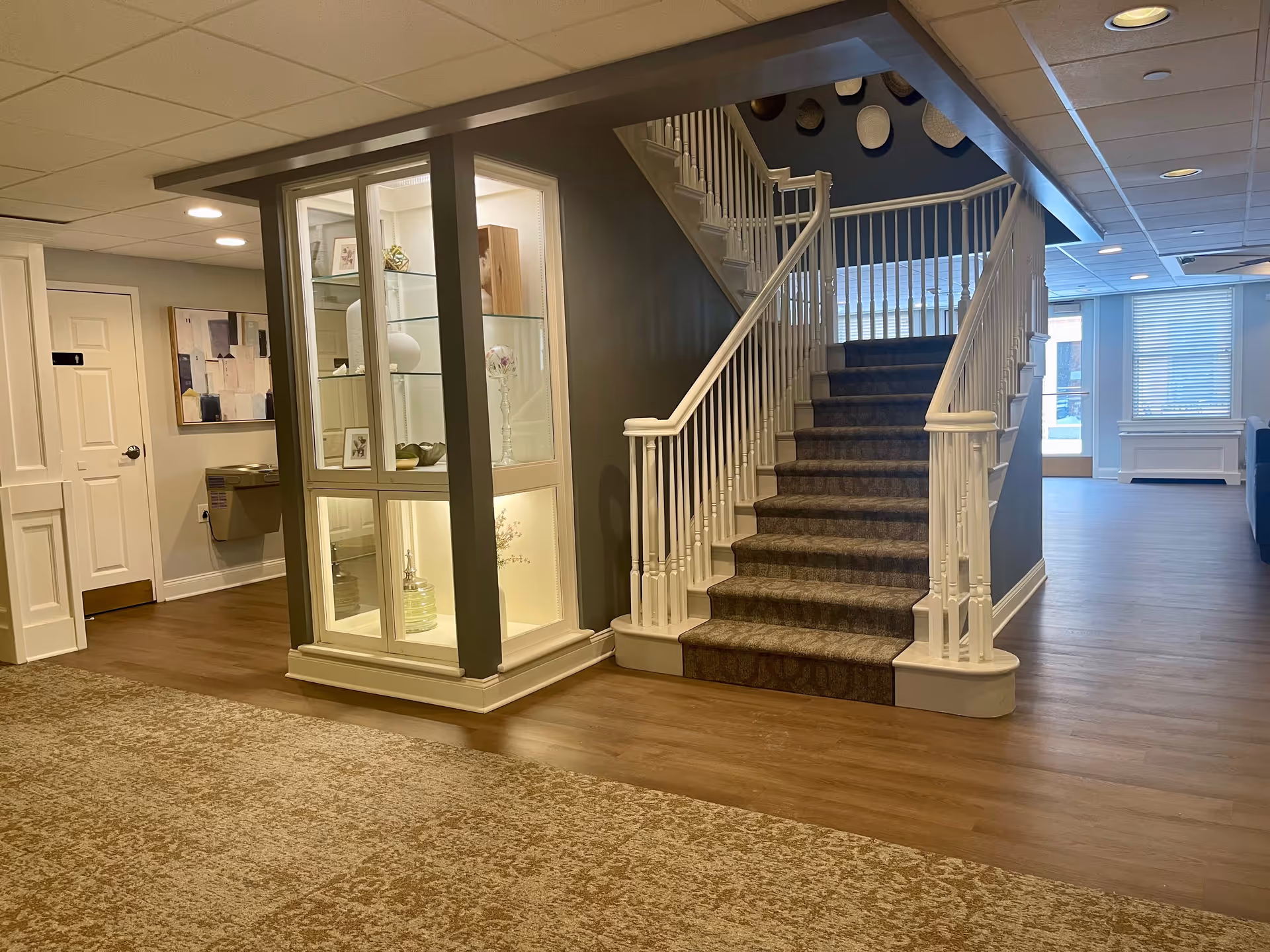 Interior view of a senior living facility showing a carpeted staircase with white railings, a display cabinet with glass shelves and decorative items, a water fountain, a door labeled with the number 1, and a hallway with wood flooring and carpeted areas.
