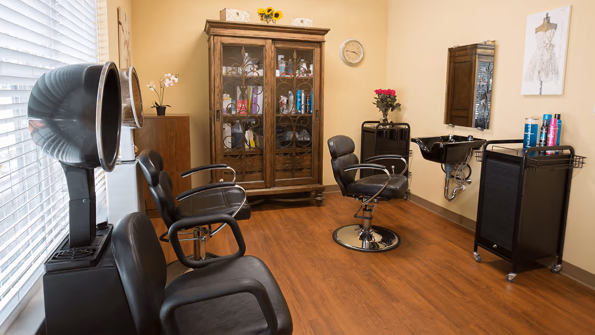 Interior of a senior living facility hair salon with two black salon chairs, two hair dryers, a wooden cabinet with glass doors containing hair products, a wall-mounted sink with a mirror above it, and a small rolling cart with hair care products. The room has wooden flooring and beige walls.