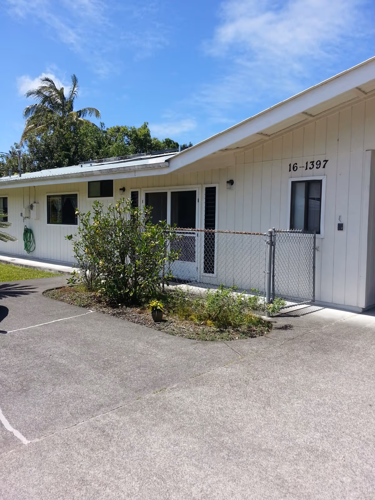 Exterior view of a single-story white building with a slanted roof, a small fenced area with a gate, and some plants and bushes in front. The building has several windows and a door, with the number 16-1397 displayed near one window. There are palm trees and a clear blue sky in the background.