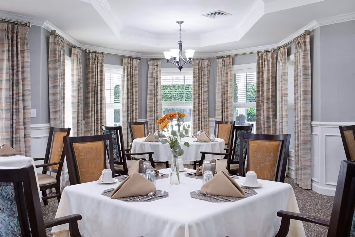 Bright dining room with tables set with white tablecloths, folded napkins, place settings and a vase of flowers, surrounded by chairs and large windows with patterned curtains.