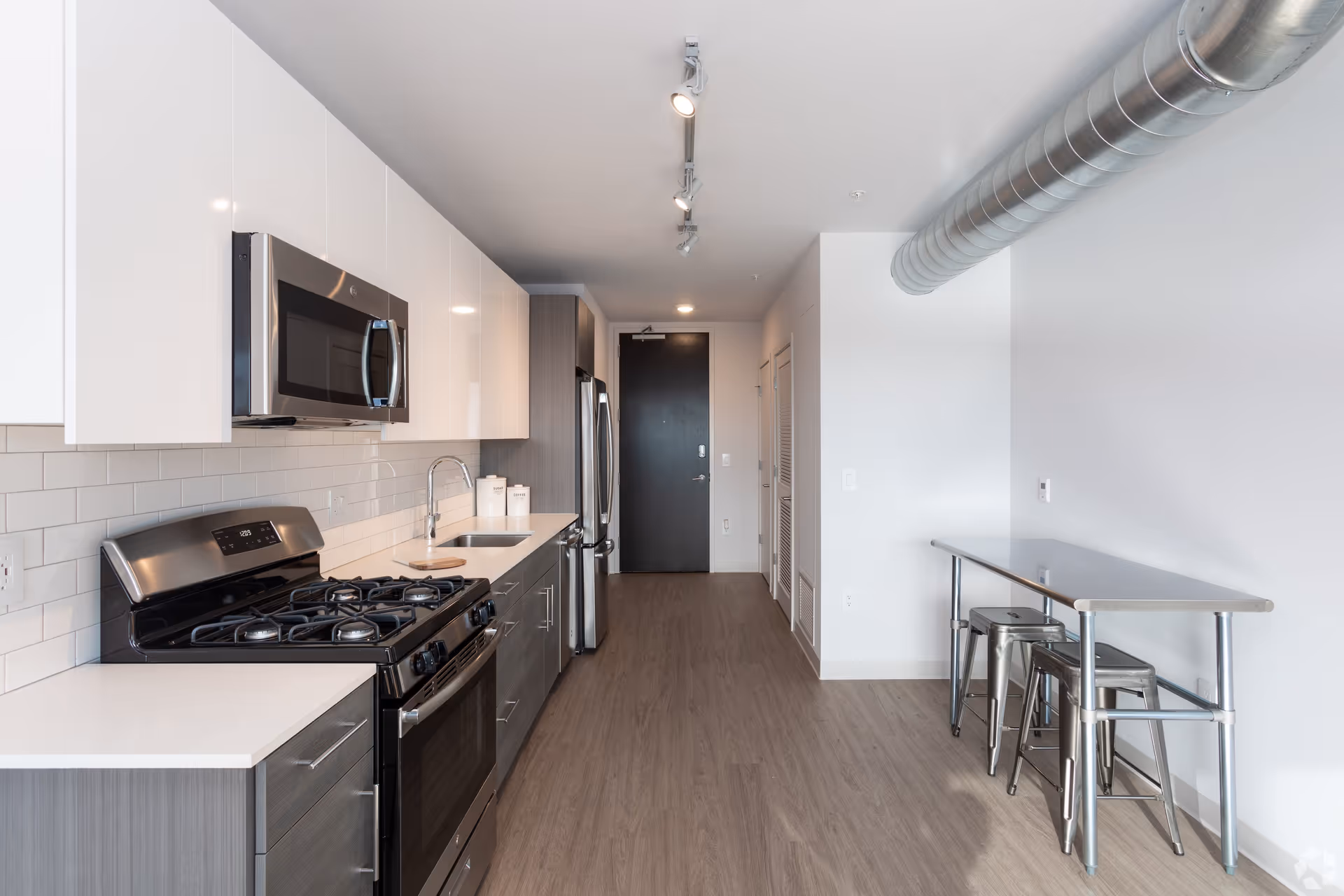 Modern kitchen with stainless steel appliances including a gas stove, microwave, and refrigerator. White upper cabinets and gray lower cabinets with a white countertop and subway tile backsplash. A narrow metal dining table with three metal stools is positioned against the right wall. The floor is wood-style laminate, and there is an exposed metal duct running along the ceiling.