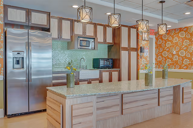 Communal kitchen area with a large island, pendant lights, stainless steel refrigerator, microwave, sink, and orange floral wallpaper.