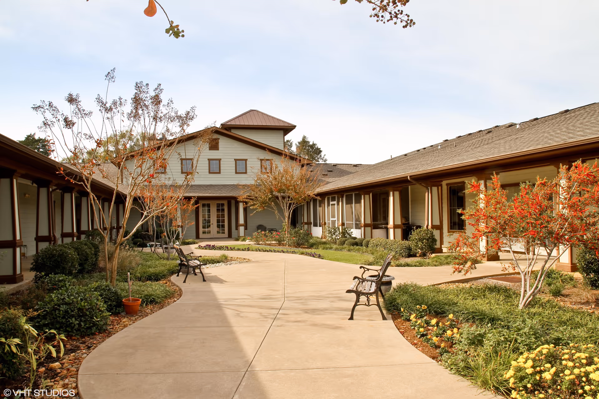 Outdoor courtyard area of Magnolia Court Assisted Living and Memory Care featuring a paved walkway, benches, landscaped garden beds with shrubs and small trees, and a building with multiple windows and a peaked roof in the background under a clear sky.