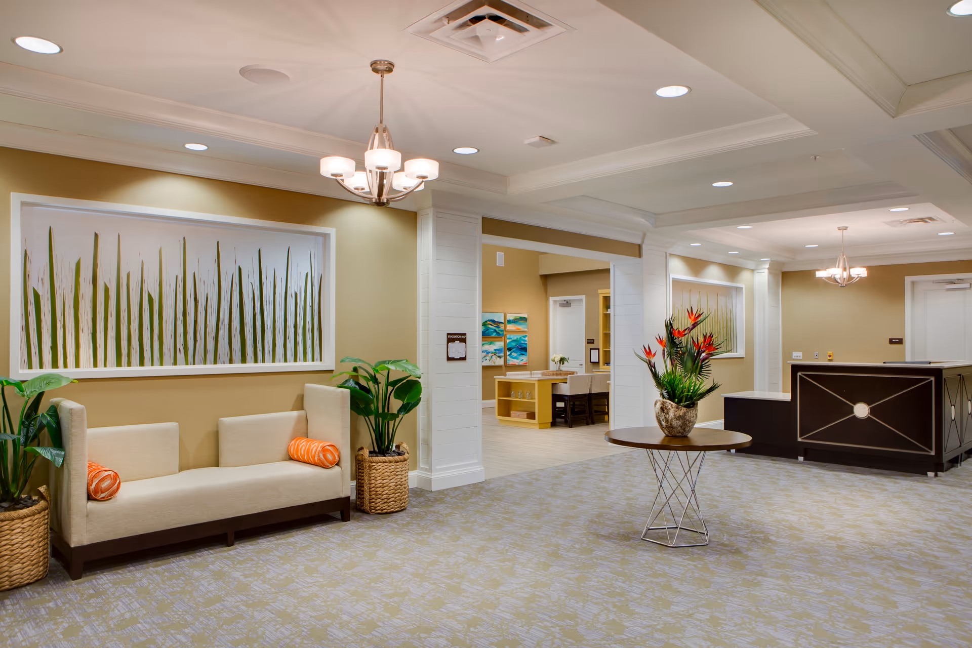 A spacious and well-lit interior common area with a beige sofa adorned with two orange cylindrical pillows, two large potted plants in woven baskets on either side of the sofa, a round table with a decorative plant arrangement in the center, and a dark reception desk in the background. The walls are painted light beige with framed artwork featuring tall green grass. The ceiling has recessed lighting and a chandelier.