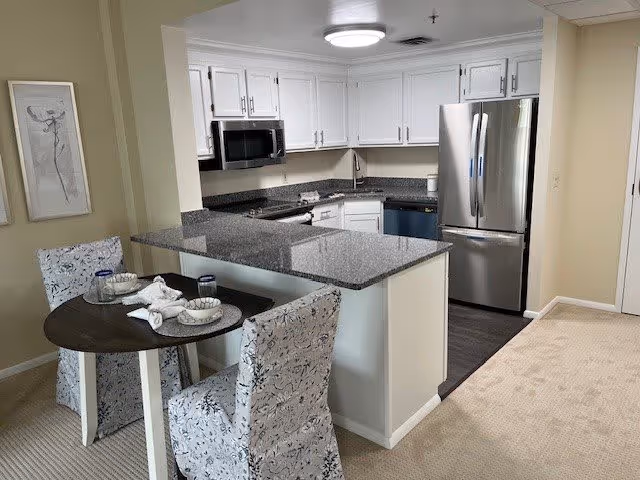 A modern kitchen and dining area with white cabinets, granite countertops, stainless steel refrigerator, microwave, and dishwasher. The dining area has a small round table with two floral-patterned chairs, set with bowls, plates, and glasses. The floor transitions from carpet in the dining area to dark wood in the kitchen. Two framed artworks hang on the wall beside the dining table.