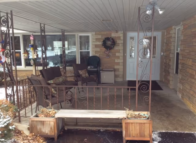 Covered front porch with seating, decorative iron railing, planters and a wreath at the entrance door.