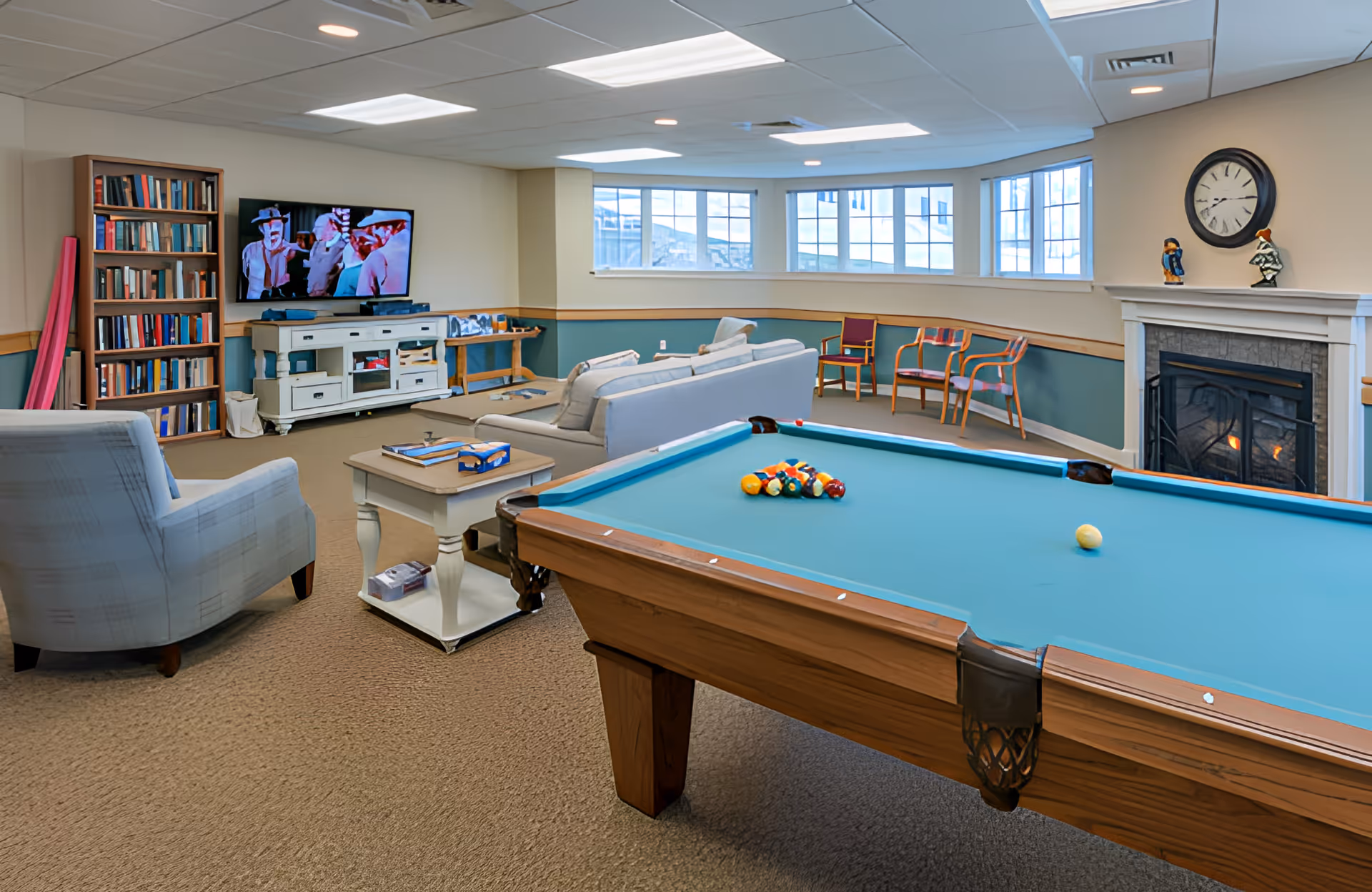 A cozy senior living common room featuring a pool table with balls set up for a game, a fireplace with a clock and decorative figurines on the mantel, a TV mounted on the wall showing a scene from a movie, a bookshelf filled with books, several chairs and sofas arranged for seating, and large windows letting in natural light.