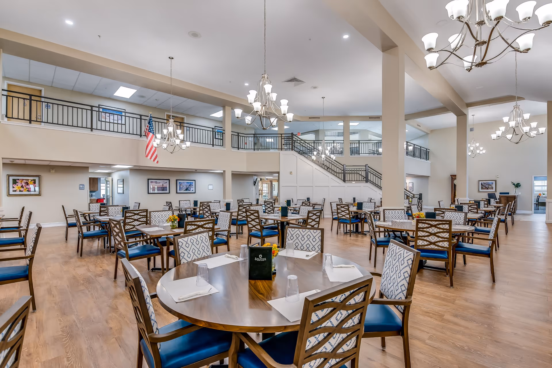 A spacious dining room with multiple round and square wooden tables surrounded by cushioned chairs with patterned backs and blue seats. The room features high ceilings with several chandeliers, a wooden floor, and a second-floor balcony with railings. An American flag hangs from the ceiling, and framed pictures adorn the walls.