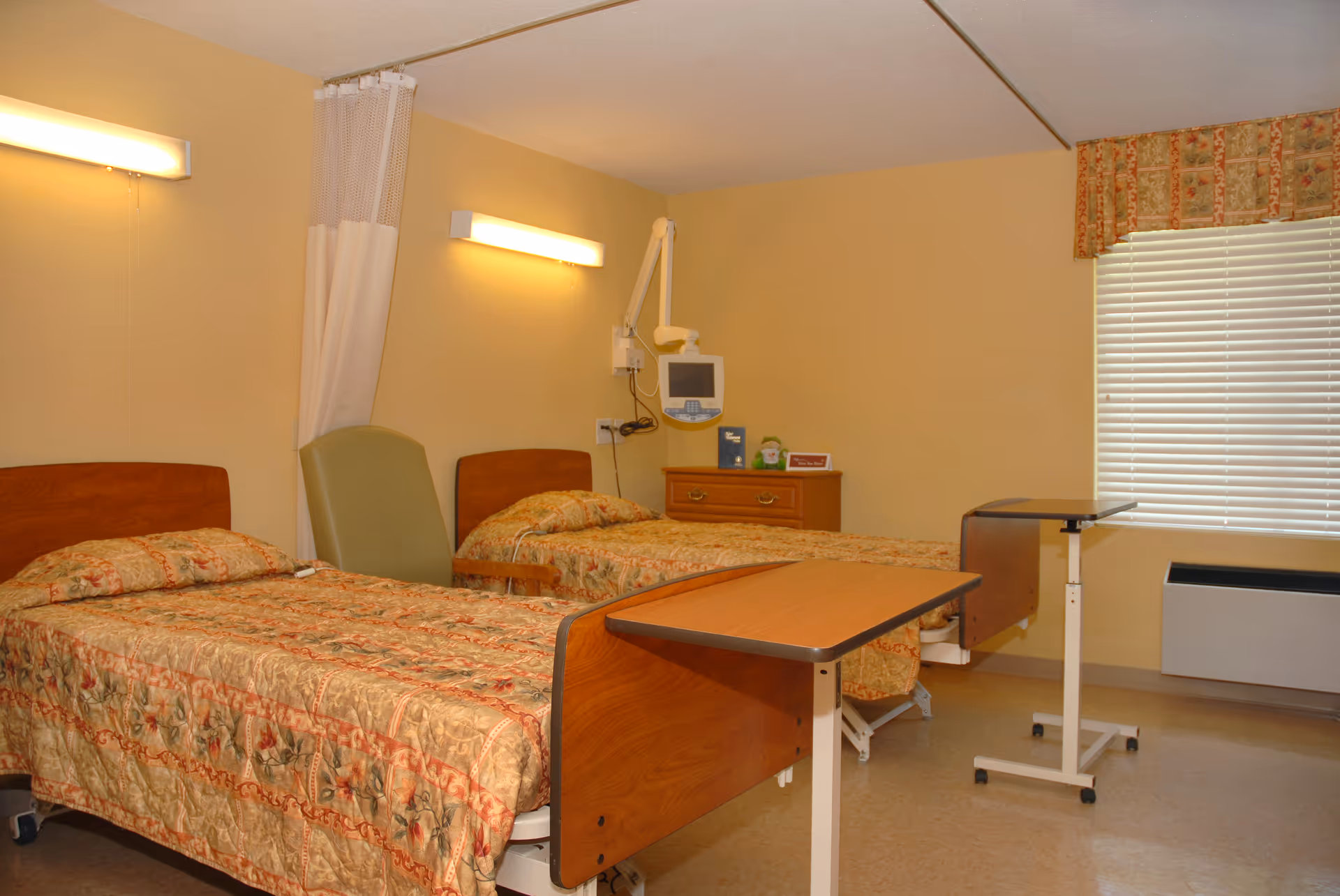 A room with two hospital-style beds, each with floral patterned bedspreads. There is a green chair between the beds, a wooden dresser with a monitor mounted on the wall above it, and two overbed tables in front of each bed. A window with blinds and a floral valance is on the right wall. The walls are painted beige and the floor is a light-colored linoleum.