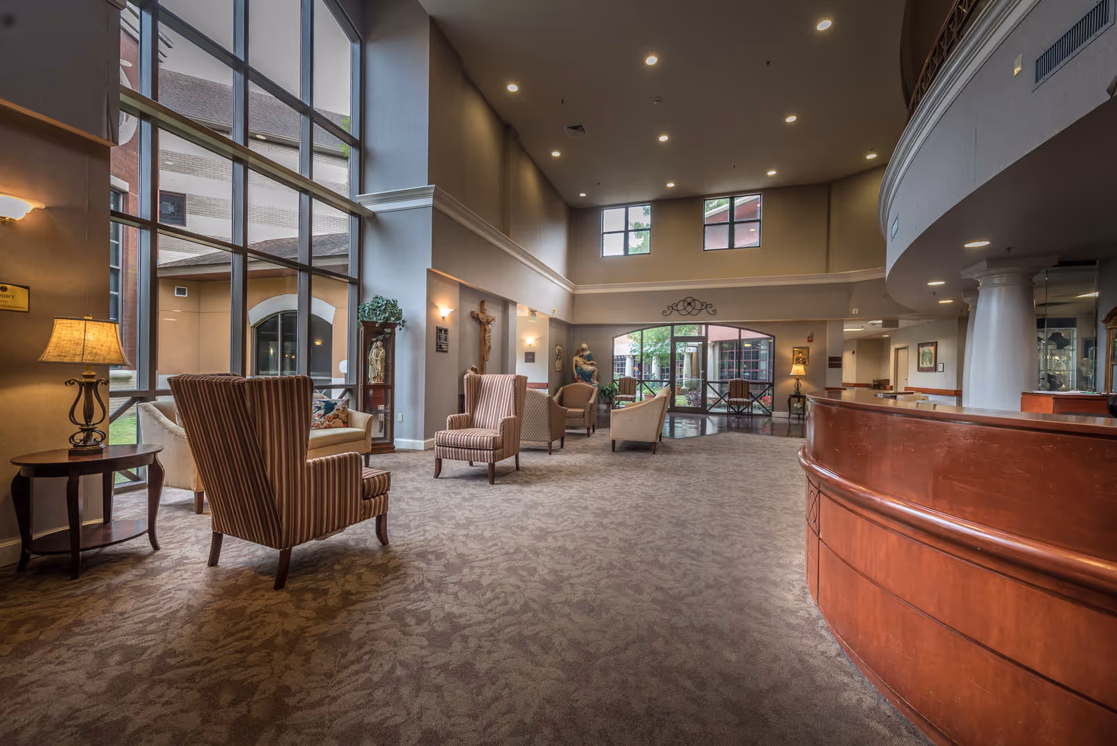 Spacious senior living lobby with high windows, multiple upholstered chairs and a curved wooden reception desk.