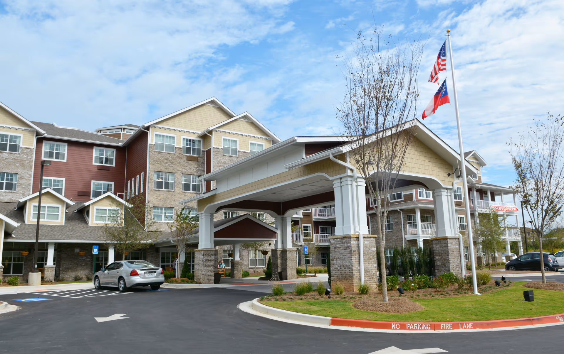 Exterior view of a multi-story senior living facility with a covered entrance supported by large white columns and brick bases. The building features a combination of brick and siding with multiple windows. There are two flagpoles with the American flag and another flag. A silver car is parked near the entrance, and the surrounding area includes landscaped greenery and a paved driveway with a 'No Parking Fire Lane' curb marking.