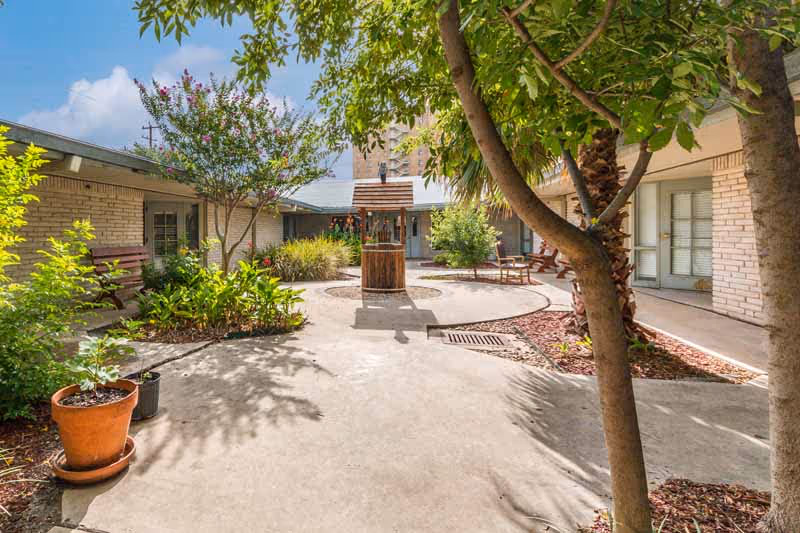 Outdoor courtyard area at Memorial Medical Nursing Center with a circular concrete pathway, a wooden wishing well in the center, surrounded by trees, plants, benches, and a building with windows and doors.