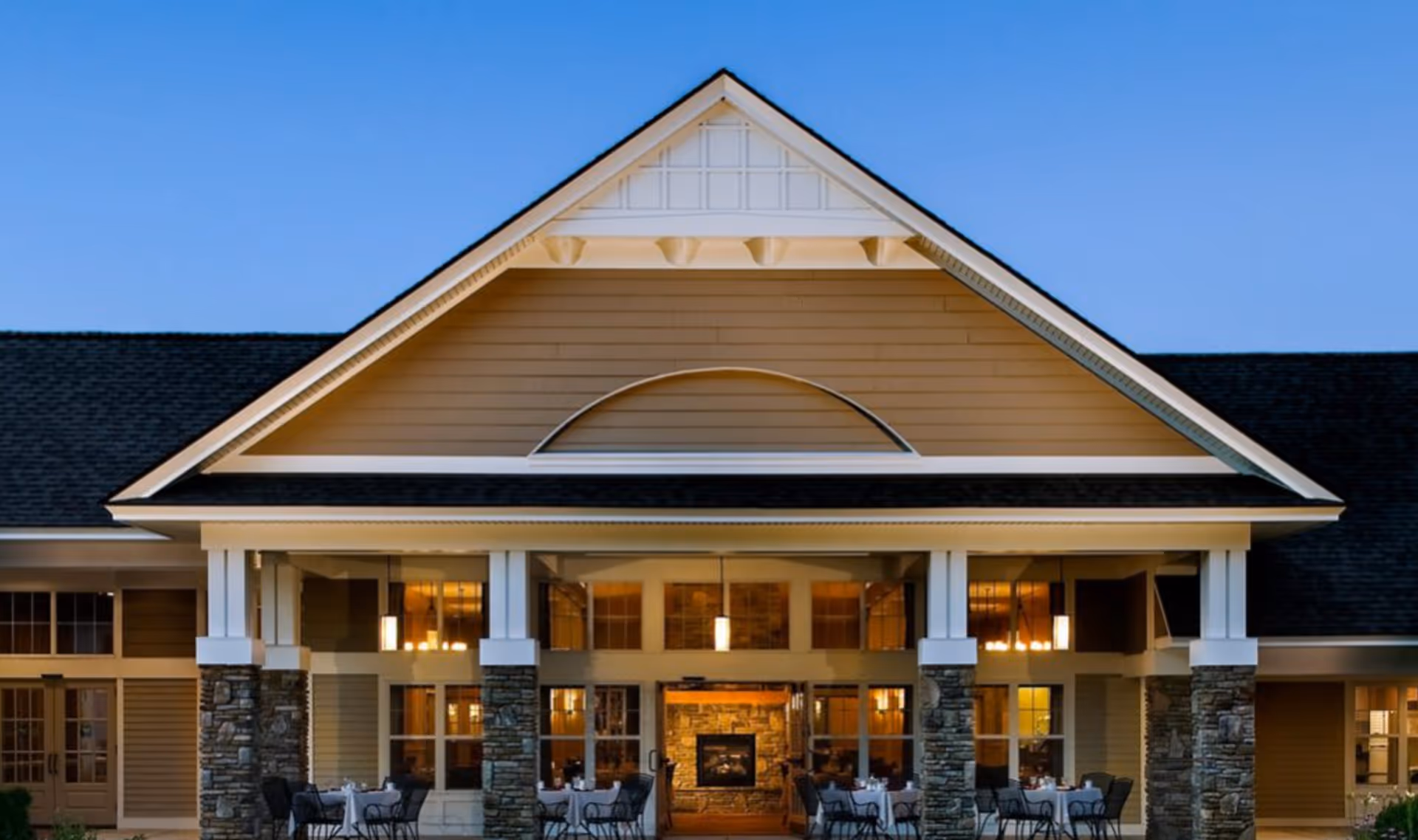 Exterior view of a senior living facility building with a peaked roof, stone pillars, and large windows. Outdoor seating with tables and chairs is visible in front of the entrance, and warm interior lighting can be seen through the windows.