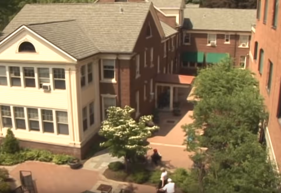 Aerial view of an outdoor courtyard area at Rochester Presbyterian Home (The Homestead) showing brick buildings with multiple windows, a paved walkway, green trees, and a few people walking and sitting in the courtyard.