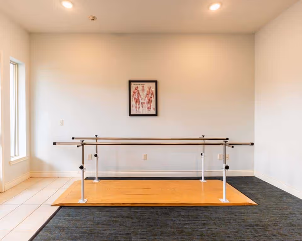 A small rehabilitation or physical therapy room with parallel bars mounted on a wooden platform, a framed anatomical poster on the wall, and a window letting in natural light.