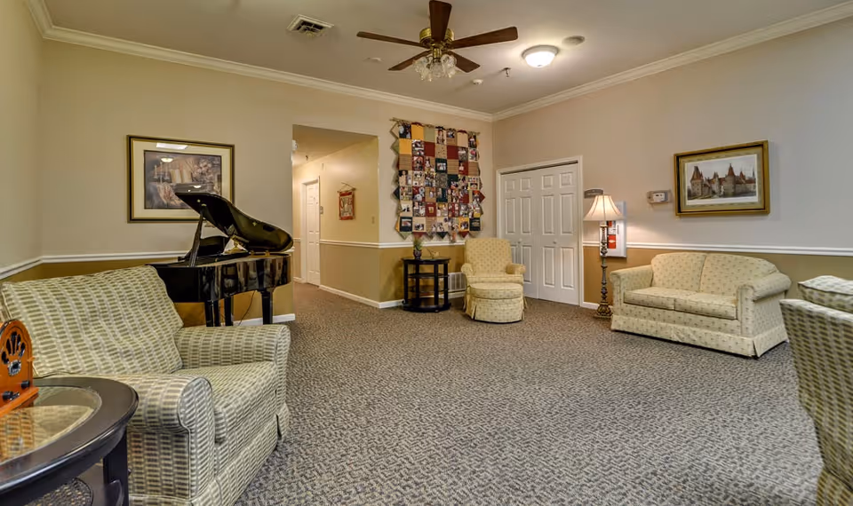 A cozy senior living facility common area with patterned carpet, beige walls, and crown molding. The room features a black grand piano, several upholstered armchairs and sofas in light patterned fabric, a standing lamp, framed artwork on the walls, a ceiling fan with lights, and a quilt or photo collage hanging on the far wall near a hallway entrance.