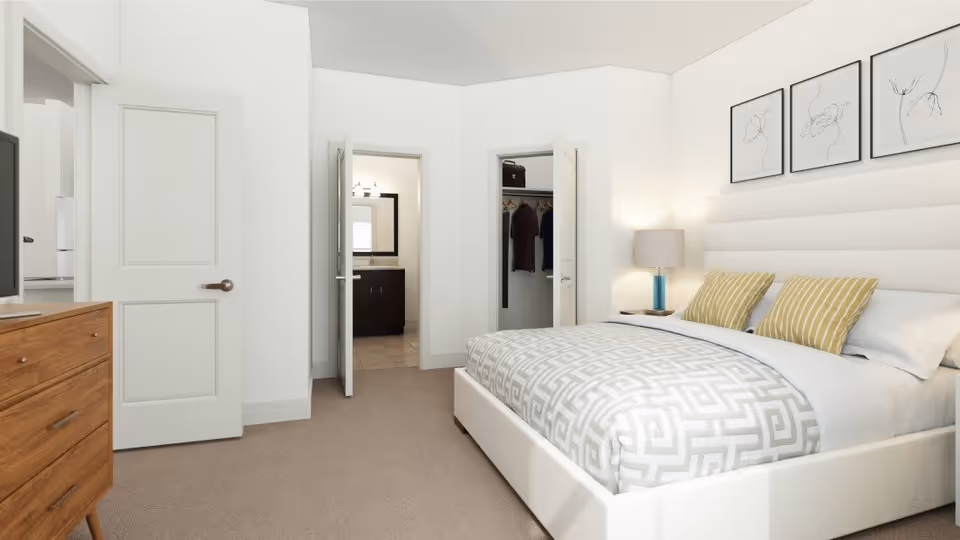 A modern bedroom with a large bed featuring a white and gray geometric patterned comforter and two yellow striped pillows. There is a wooden dresser with a TV on top to the left, a bedside table with a lamp on the right, and three minimalist framed artworks on the wall above the bed. Two open doors reveal a bathroom with a vanity and a closet with hanging clothes.