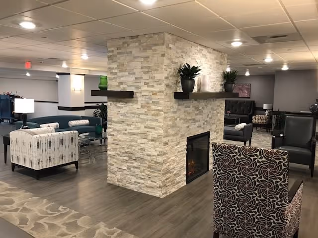 A spacious senior living facility common area with a central stone fireplace. The room features various seating options including patterned armchairs, a teal sofa, and cushioned chairs arranged in small groupings. The floor is a combination of wood and carpet, and the ceiling has recessed lighting. Decorative plants and shelves adorn the fireplace, creating a cozy and inviting atmosphere.