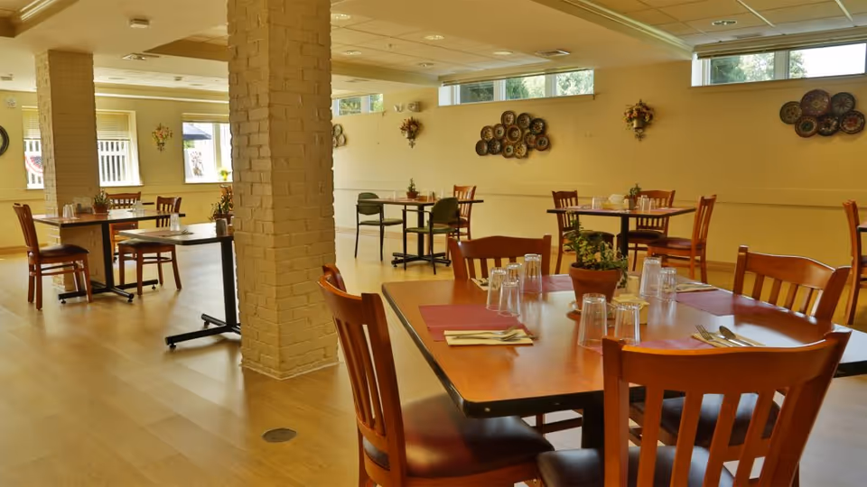 A bright dining room in a senior living facility with several wooden tables and chairs. Each table is set with placemats, utensils, and upside-down drinking glasses. There are decorative plants on the tables and wall decorations on the light yellow walls. Large windows allow natural light to fill the room.