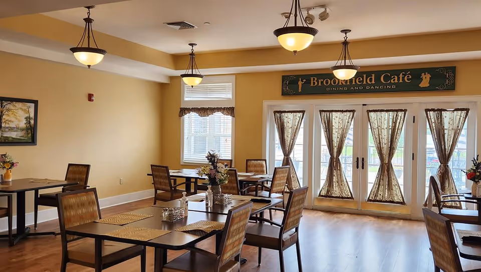 A dining area with several tables and chairs arranged neatly. The room has warm yellow walls, wooden flooring, and three hanging ceiling lights. There are windows with sheer curtains and a sign above the door that reads 'Brookfield Café Dining and Dancing'. Floral centerpieces are placed on the tables.