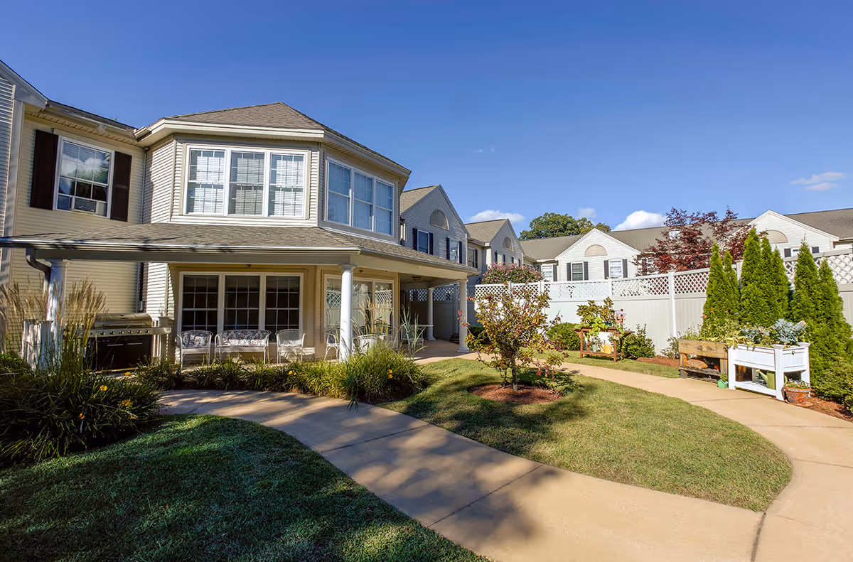 Landscaped courtyard with a paved walkway, planter boxes and patio seating alongside a two-story senior living building under a clear blue sky.