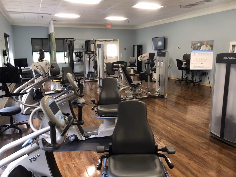 Interior view of a fitness room with various exercise machines including stationary bikes and weight machines, wooden flooring, light blue walls, and a TV mounted on the wall. There are also chairs and a poster stand in the background.