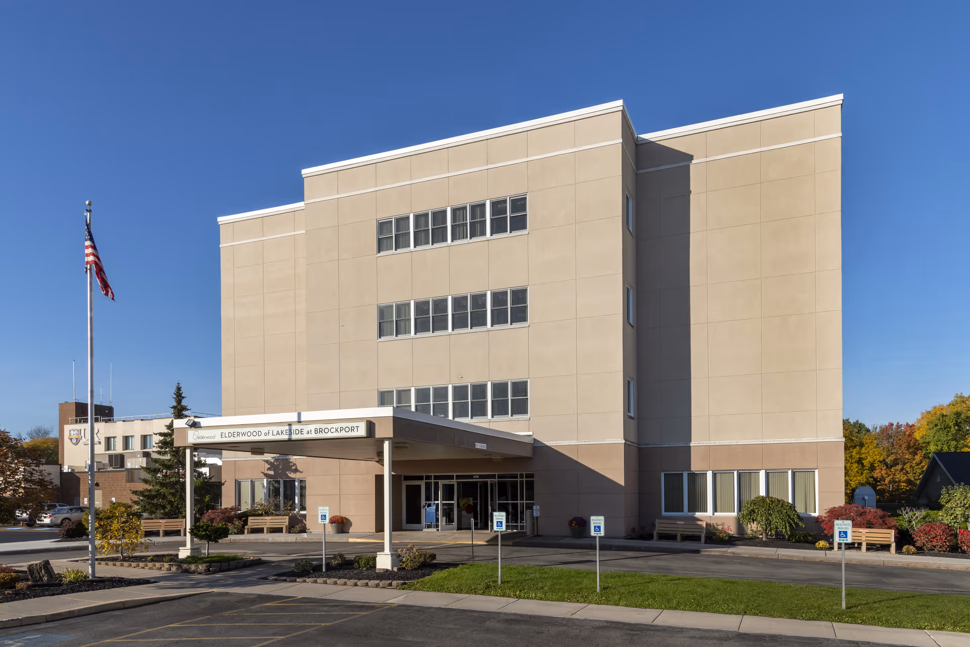 Exterior view of a multi-story beige building with a covered entrance labeled 'Elderwood of Lakeside at Brockport'. There is an American flag on a flagpole to the left, several handicap parking signs in front, and some landscaping with bushes and trees around the building under a clear blue sky.