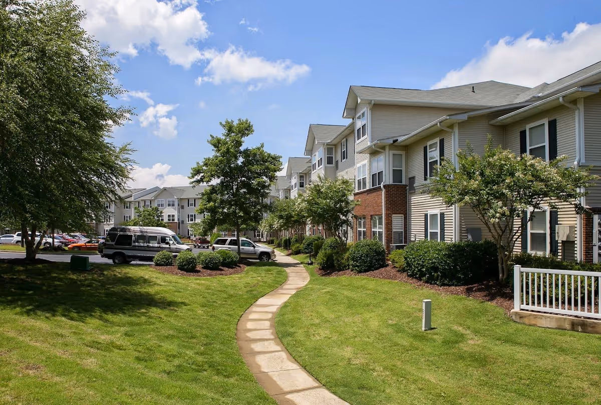 Outdoor view of a senior living facility named Lakeside Gardens showing a paved walkway through a well-maintained grassy area with bushes and trees, leading to multi-story residential buildings under a partly cloudy sky.