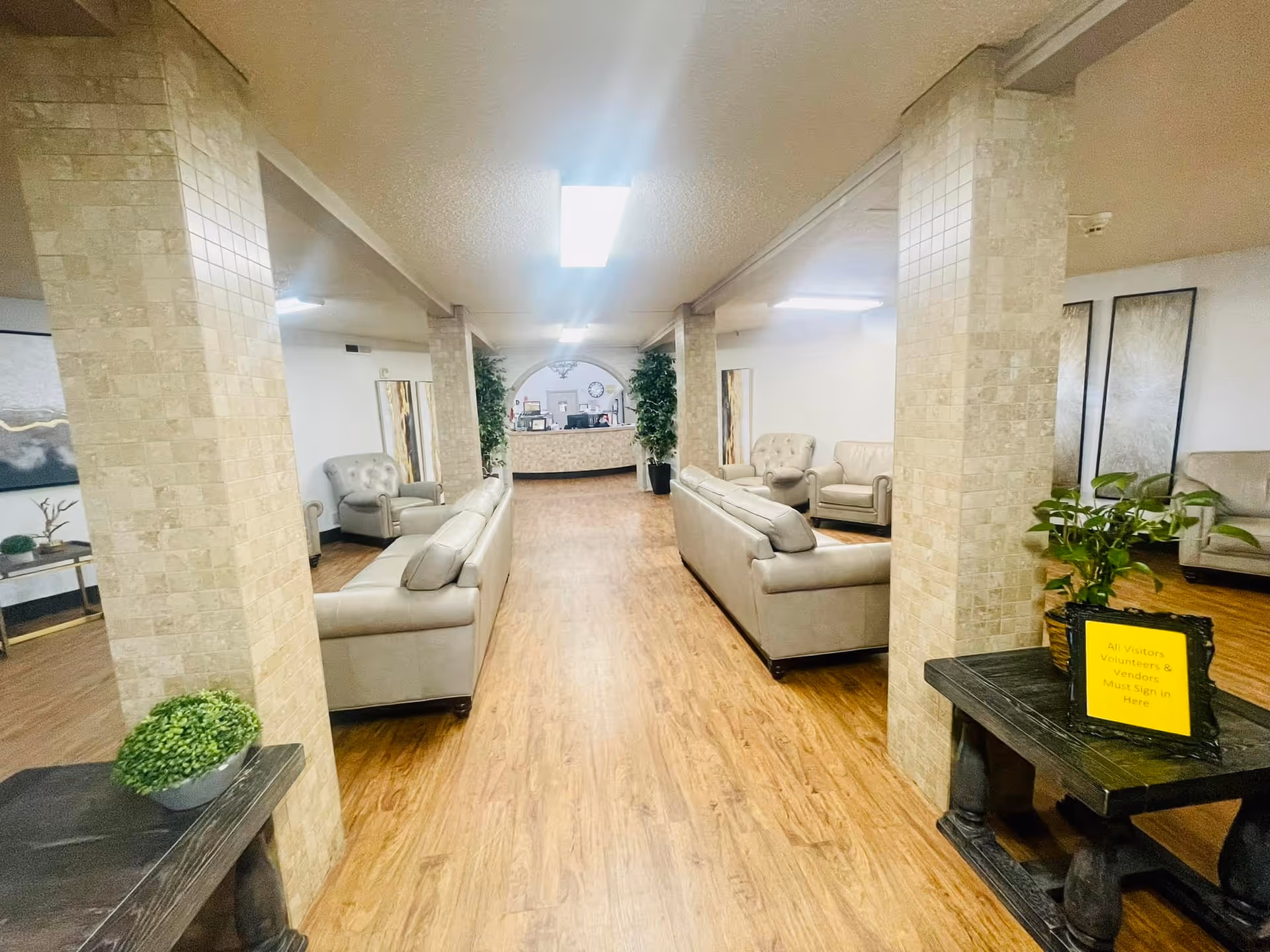 A spacious and well-lit common area with beige leather sofas and armchairs arranged along a wooden floor. The room features tiled pillars, potted plants, and a reception desk at the far end. A small table on the right holds a yellow sign that reads 'All Visitors Volunteers & Vendors Must Sign In Here.'