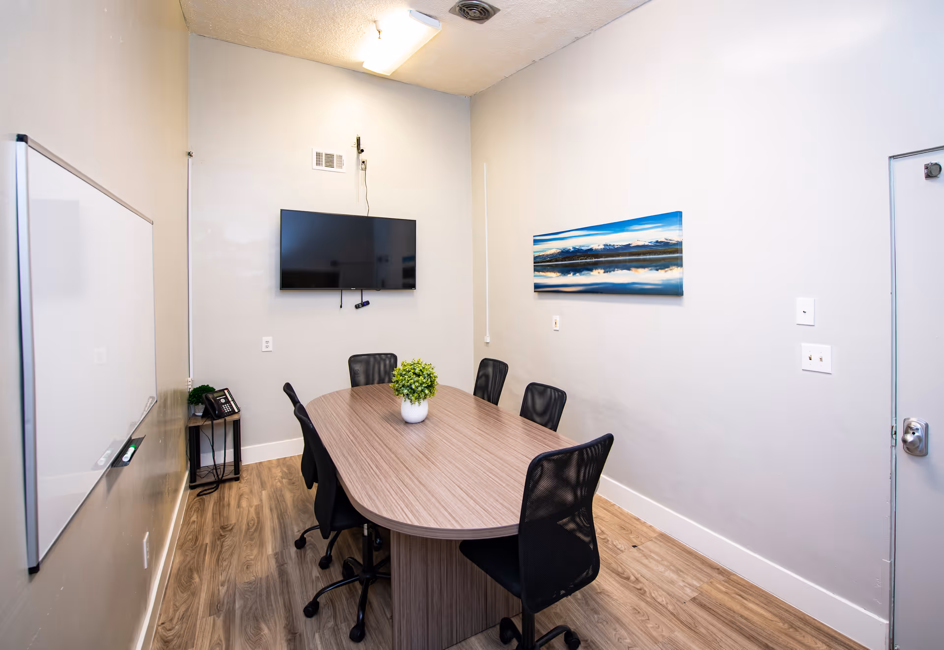 A small conference room with a wooden oval table surrounded by six black mesh office chairs. A flat-screen TV is mounted on the wall opposite the door. There is a whiteboard on the left wall and a small side table with a phone and a plant in the corner. A landscape photo is hung on the right wall. The room has light-colored walls and wood flooring.