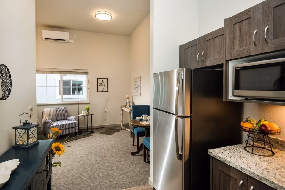 Studio-style apartment interior showing a compact kitchen with stainless steel refrigerator and microwave, and a cozy living area with a striped armchair and small dining table.
