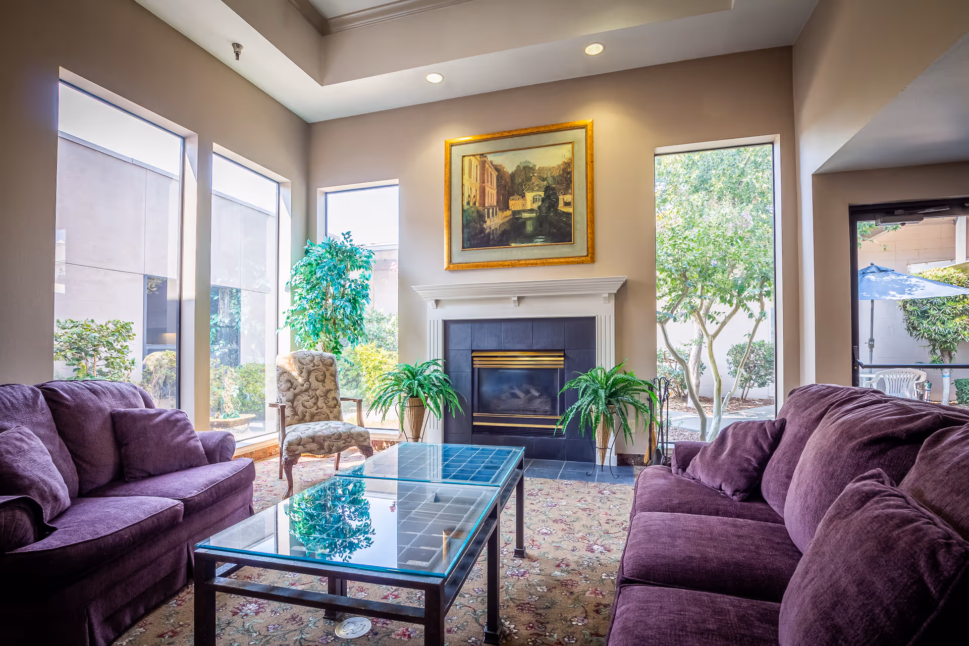 A cozy living room with two purple sofas facing each other, a glass coffee table in the center, a patterned armchair, and a fireplace with a framed painting above it. Large windows let in natural light and show greenery outside.
