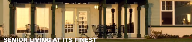 Exterior view of a senior living facility at dusk with large windows and a patio area furnished with tables and chairs.