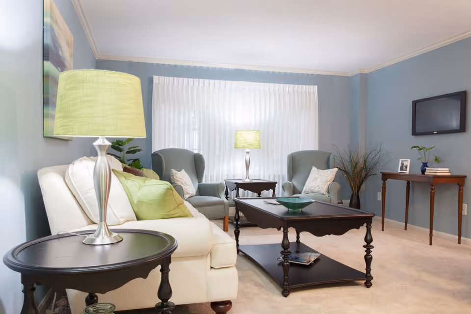 Light-filled living room with a cream sofa, two blue-green armchairs, dark wooden coffee and side tables, green lamps, and a wall-mounted TV.