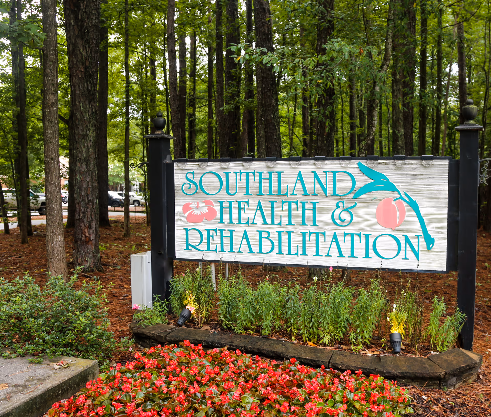 A wooden sign for Southland Health & Rehabilitation surrounded by greenery and red flowers, with tall trees in the background.