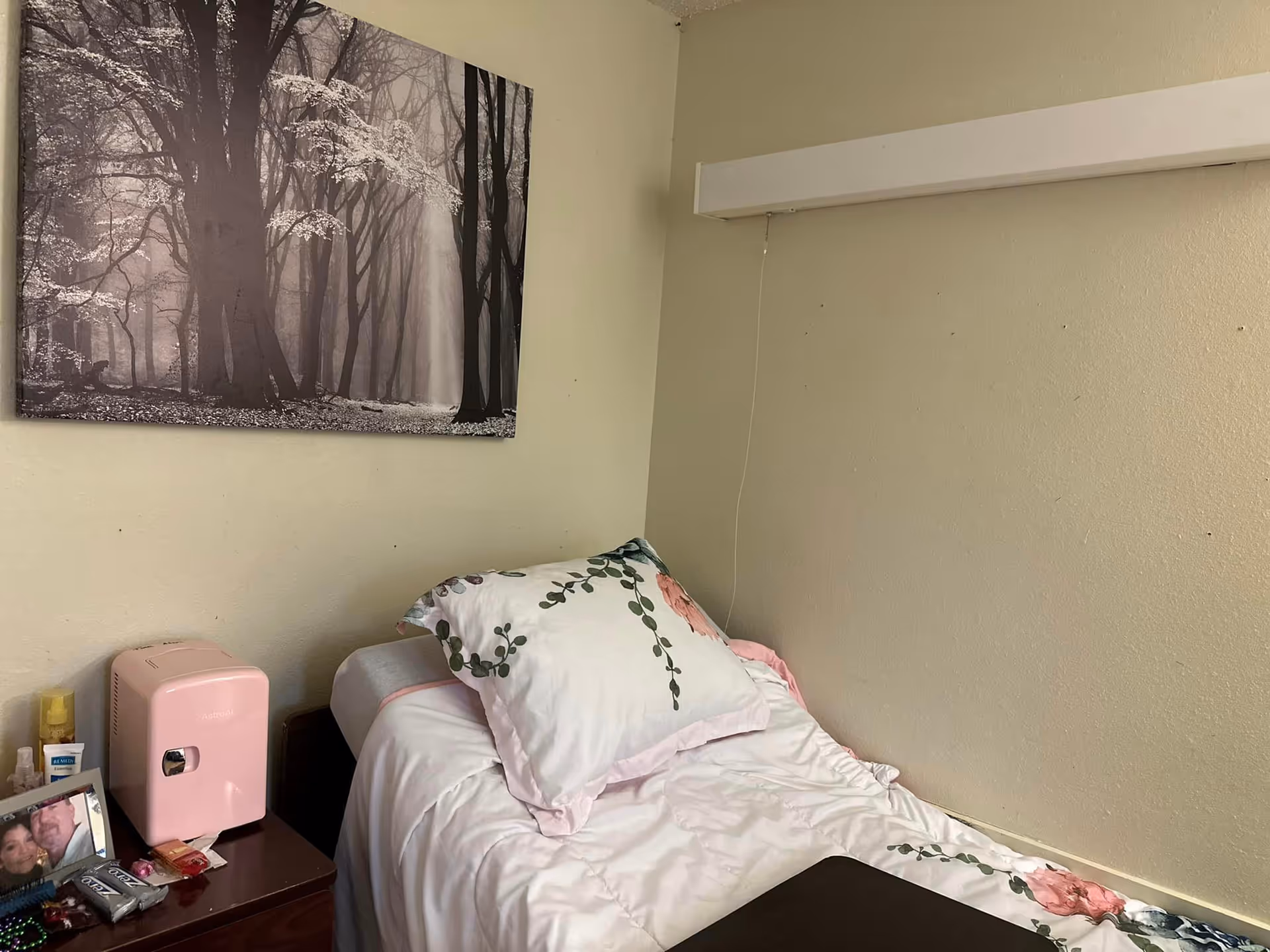 A small bedroom with a single bed featuring white bedding with floral patterns. Next to the bed is a wooden nightstand holding a pink mini fridge, a framed photo, and various small items. Above the bed hangs a black and white picture of a forest with tall trees.