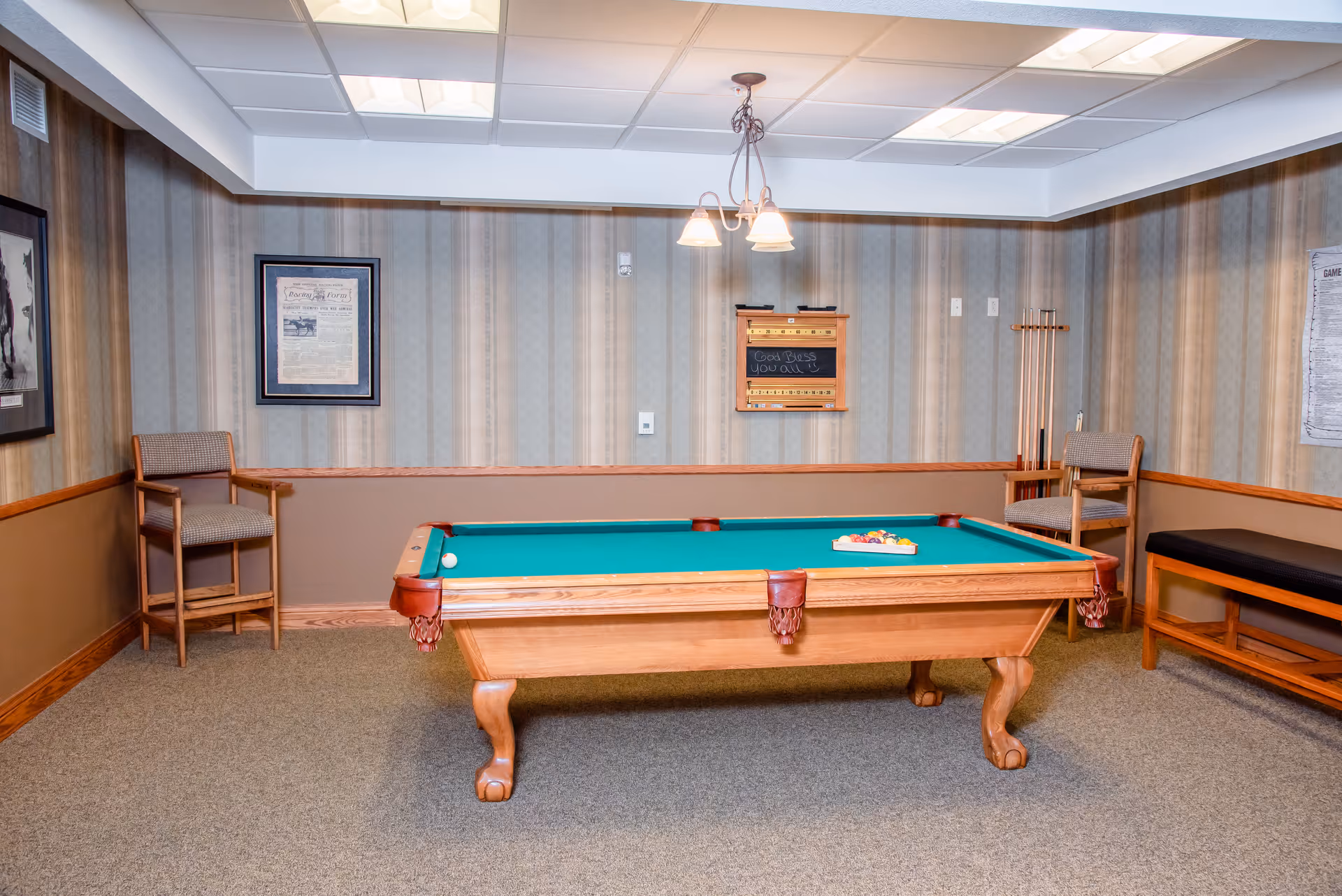 A carpeted recreation room with a wooden pool table, cue rack, chairs, and framed wall decor.