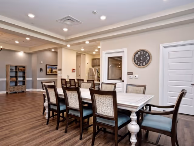 A spacious dining area in a senior living facility featuring a long white dining table with eight cushioned chairs. The room has wood flooring, beige walls, recessed ceiling lights, a wall clock, and a door leading to another room. In the background, there is a kitchen area with stainless steel appliances and a wooden shelving unit against the far wall.