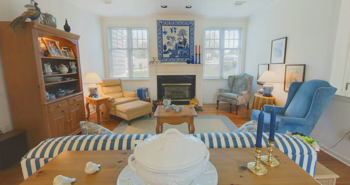 A cozy living room with a fireplace centered between two windows. The room features a beige armchair with an ottoman, a blue patterned armchair, and a blue wingback chair. A wooden coffee table with a tea set sits on a light-colored rug. There is a wooden cabinet with decorative items and framed photos on the left, and two table lamps on either side of the room. Blue candles in brass holders are on a wooden surface in the foreground.