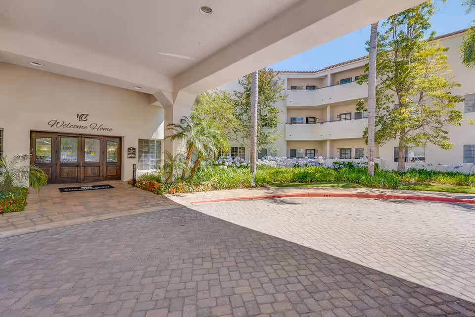 Entrance area of Meridian at Anaheim Hills facility with a covered driveway, potted plants, and a welcoming sign that reads 'Welcome Home' above wooden double doors. The building exterior is light-colored with balconies and surrounded by greenery and palm trees under a clear blue sky.