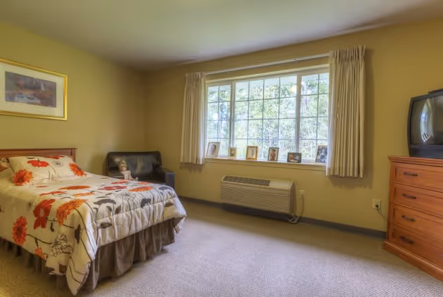 A bedroom with a single bed covered in a floral bedspread, a black armchair in the corner, a large window with beige curtains, a wooden dresser with an old-style TV on top, and several framed photos on the windowsill.