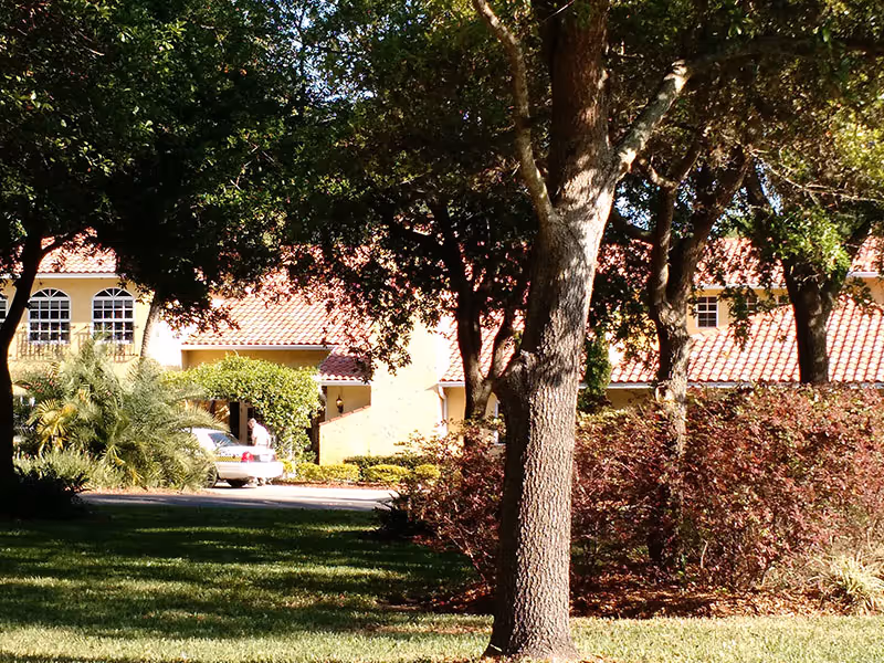 View of a senior living facility building partially obscured by trees and bushes, with a person walking near a parked car in front of the building. The building has a light yellow exterior and a red tile roof.