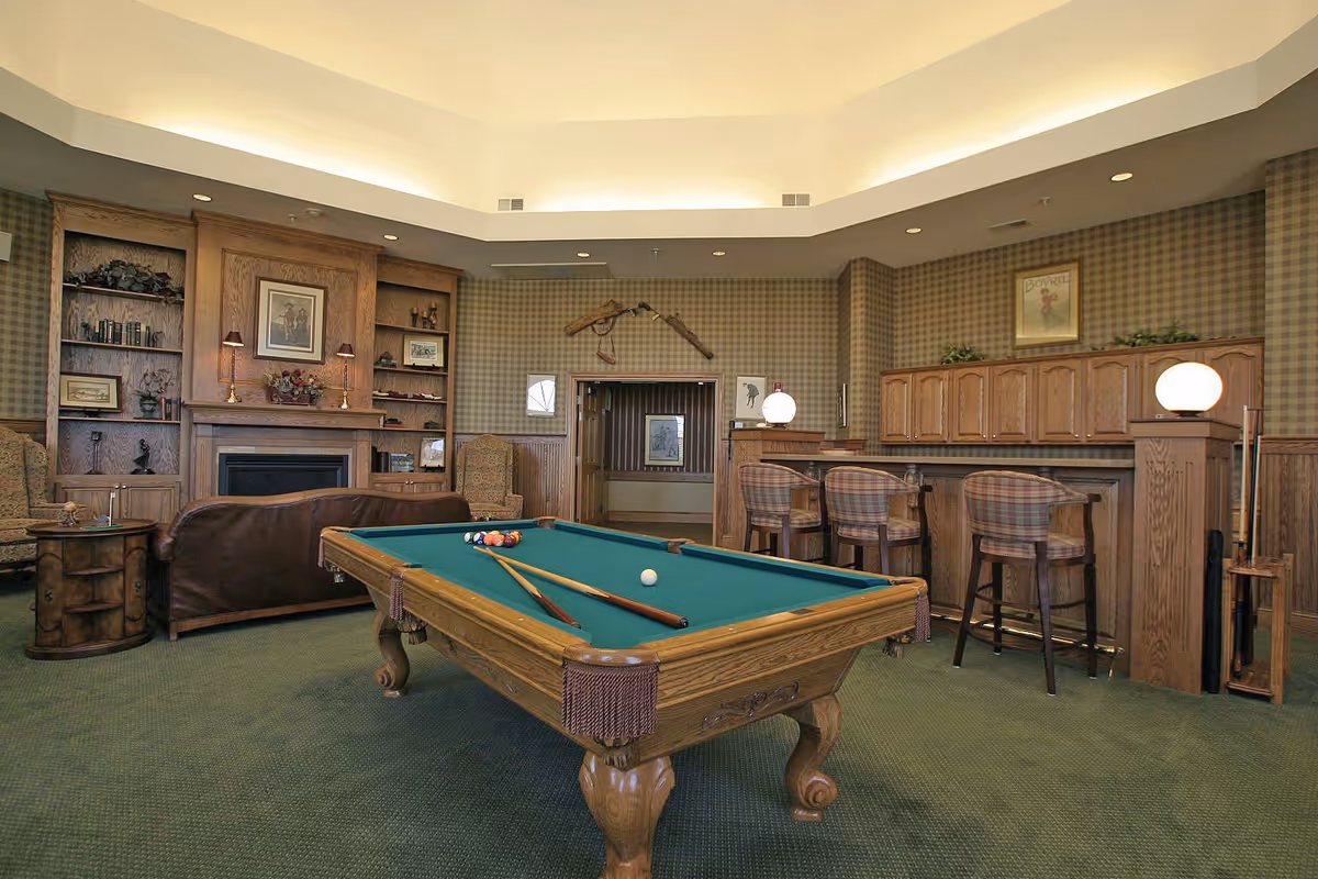 A cozy lounge with a pool table in the foreground, leather seating, built-in shelves, and a bar with stools.