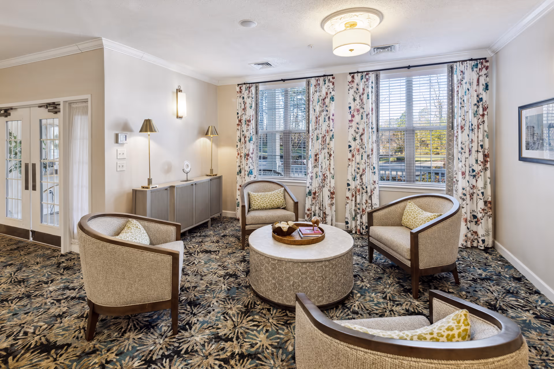 Sunlit common sitting area with four upholstered armchairs around a round coffee table, floral curtains and patterned carpet.