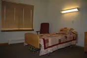 A bedroom in a senior living facility with a bed, quilt, window blinds, and chair.