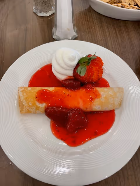 A plate with a rolled crepe topped in strawberry sauce, a sliced strawberry and a dollop of whipped cream on a wooden table.