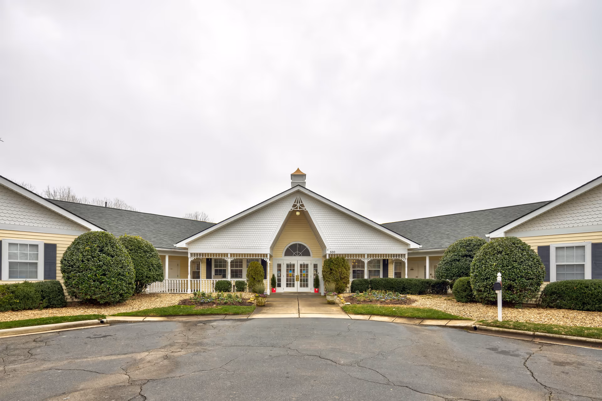 Front entrance of a single-story senior living building with a central covered porch, symmetrical wings, and neatly trimmed shrubs.