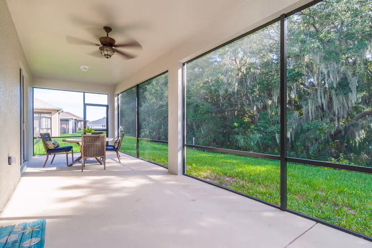 Screened-in covered patio with a ceiling fan, table and chairs overlooking a grassy yard and trees.