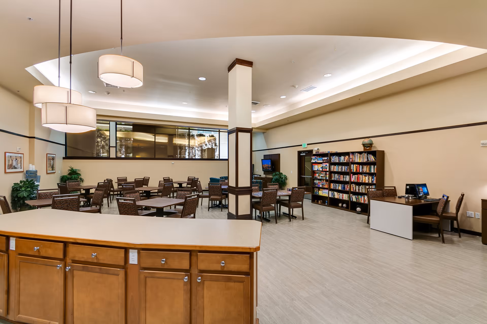 A spacious common area in a senior living facility featuring multiple tables and chairs arranged for socializing or activities. There is a bookshelf filled with books and games, a desk with a computer, and large pendant lights hanging from the ceiling. The room has light-colored walls and flooring, with a partial view of a kitchen counter in the foreground.