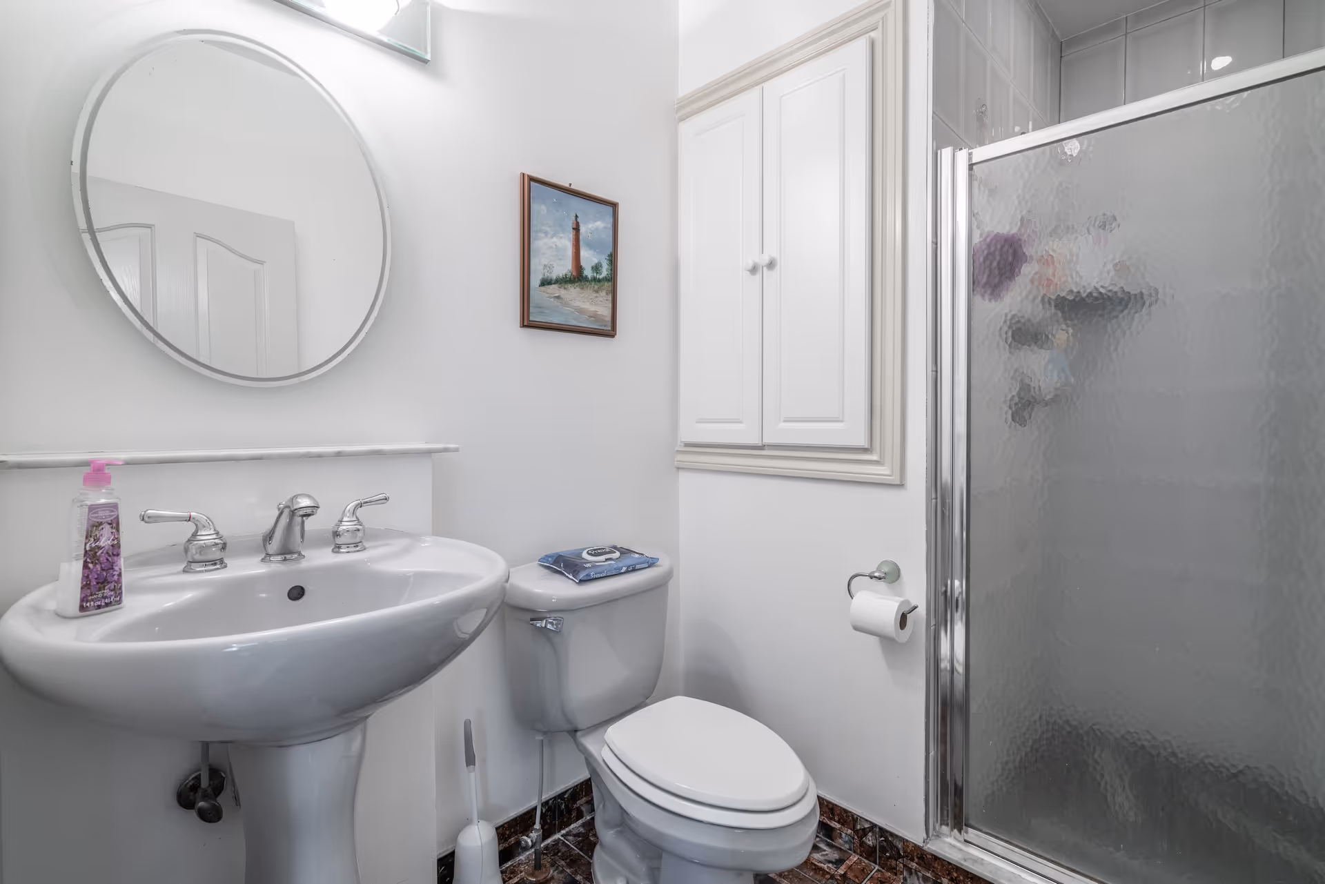 A small bathroom with a white pedestal sink, a round mirror above it, a toilet with a pack of wipes on the tank, a toilet paper holder with a roll of toilet paper, a white cabinet mounted on the wall, and a shower with a frosted glass door.
